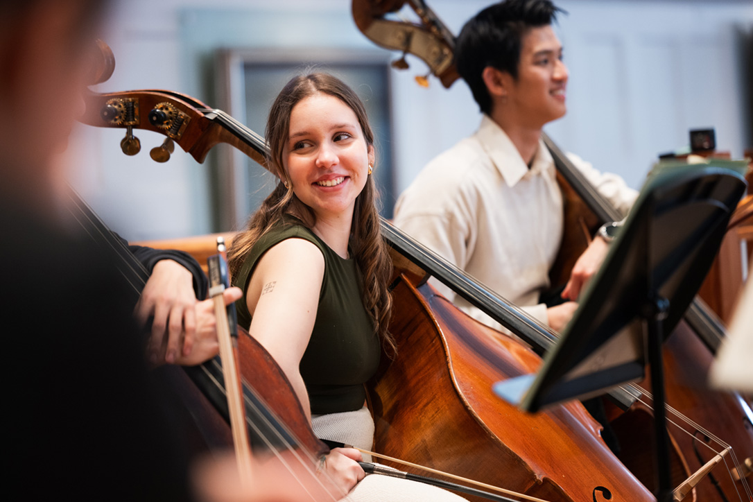 A female double bass player, her double bass leaning against her, chatting to other students, sitting down in an orchestra rehearsal.